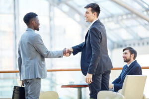 Portrait of smiling successful businessman greeting African-American man in meeting shaking hands at table in office building