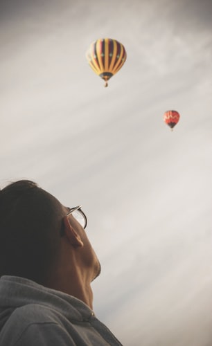 man looking up at a gray sky with hot air balloons