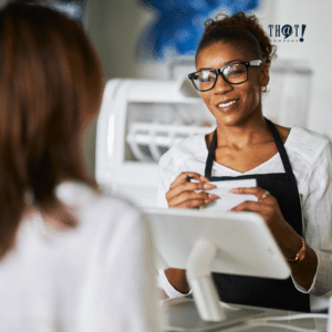 Attentiveness | A Girl In A Casher Holding A Pen And Paper Smiling To a Customer 